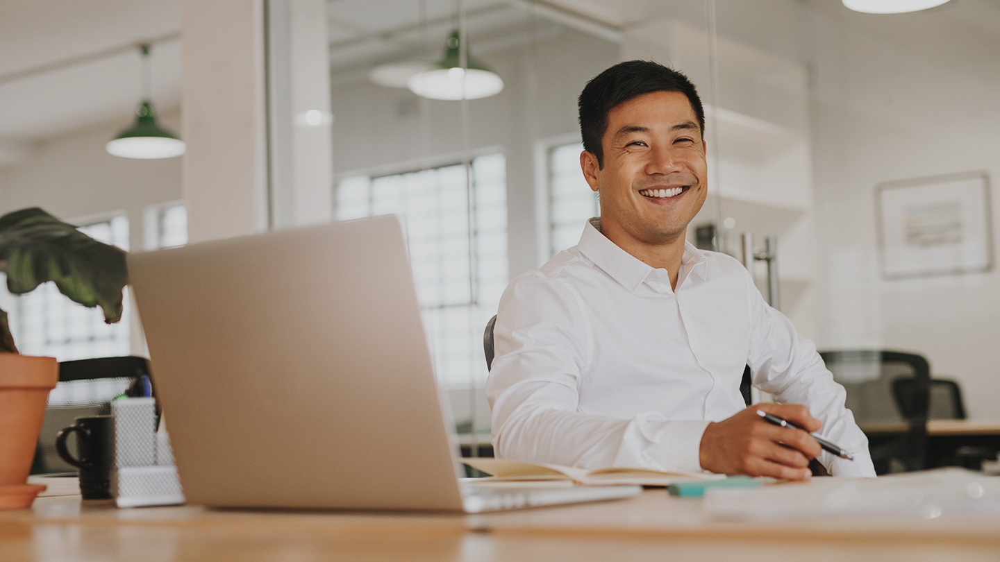 A man sits at a desk with laptop in an office setting smiling to camera